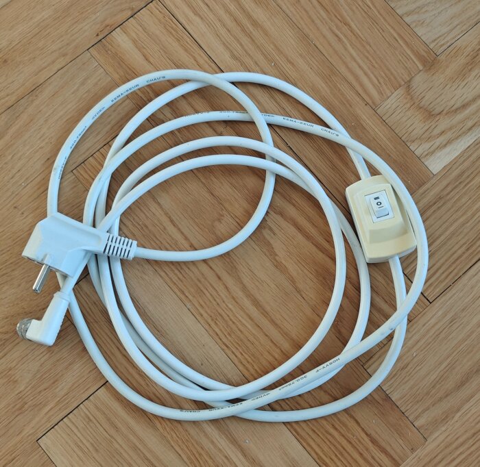 Vit kabel med strömbrytare och stickkontakt ligger på ett parkettgolv.