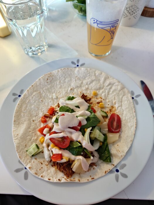 Tortilla med pulled pork toppad med sallad, tomat, gurka och sås. Glas med dryck och ett vattenglas i bakgrunden.
