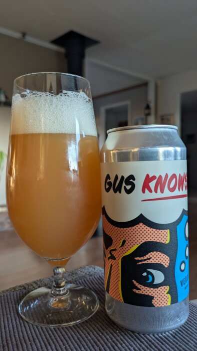 Ölglas med skummande öl bredvid en ölburk märkt "Gus Knows" från Verdant, stående på en bordsyta.