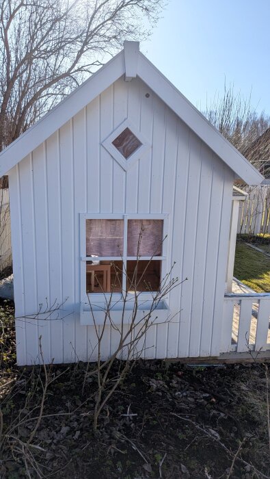 Vitt lekstuga med fönster och liten veranda, omgiven av träd och buskar i en trädgård.