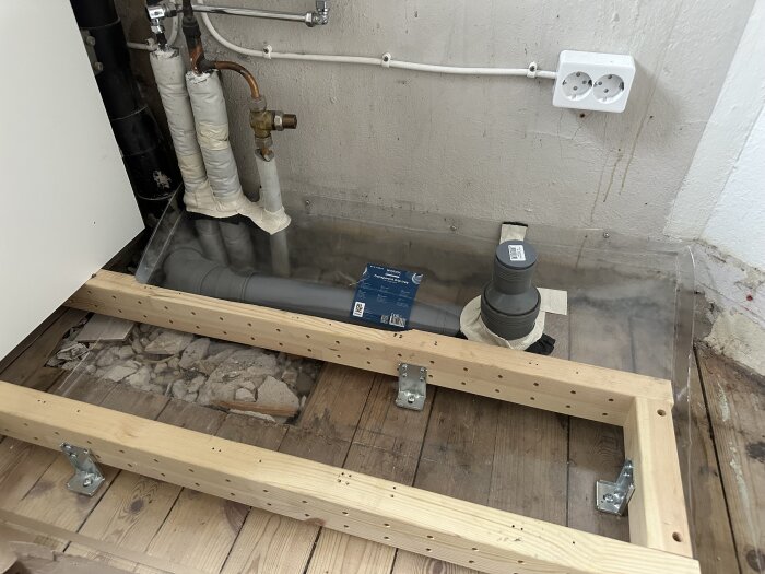 Installation av rörsystem med träram, kopparrör och ett avloppsrör. Ett eluttag syns på väggen.
