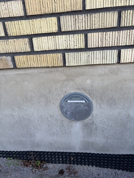 Ventilationsöppning i tegelfasad med betonggrund, omgiven av mark med grus och vegetation.