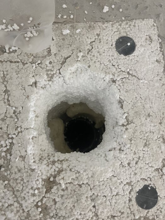 Öppen golvytan med borttagen frigolit kring en golvbrunn, omgiven av frigolitkulor och skum, redo för installation och fixering.