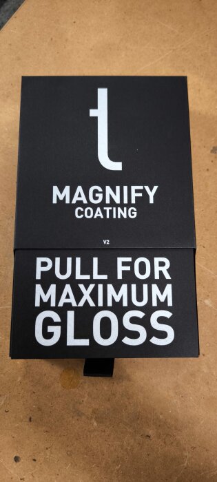 Svart förpackning med texten "Magnify Coating V2" och "Pull for Maximum Gloss" på ett träbord.