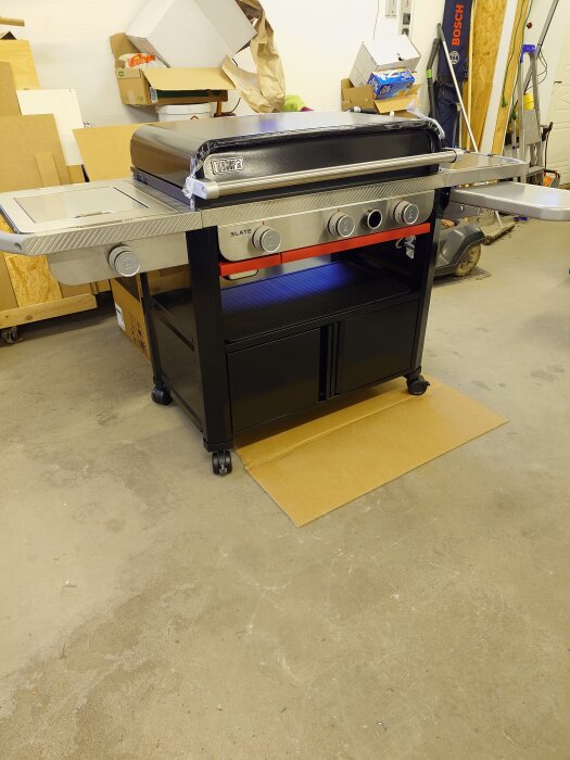 Gasolgrill av märket Weber uppställd inomhus, med förvaringshylla och sidobord, placerad på kartongbit i ett garageomgiven av kartonger och verktyg.