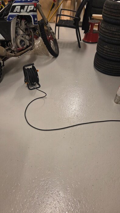 Motordel av en AJP-motorcykel i ett garage med ihoprullad elkabel på golvet och staplade däck i bakgrunden.