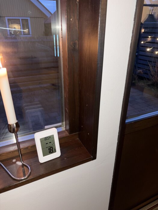 Mörka träfönstersmygar med digital termometer på fönsterbrädan, ljusstake med brinnande ljus bredvid.
