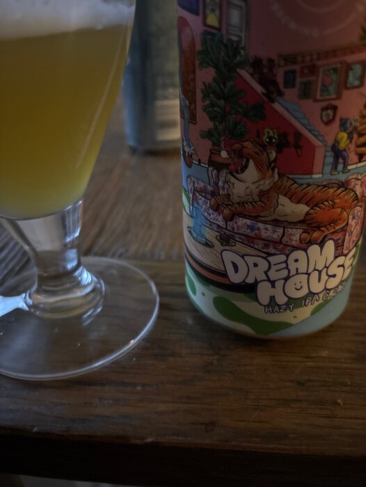 Ett glas med gul vätska bredvid en färgglad burk märkt "Dream House Hazy IPA 6.2%" på ett träbord.