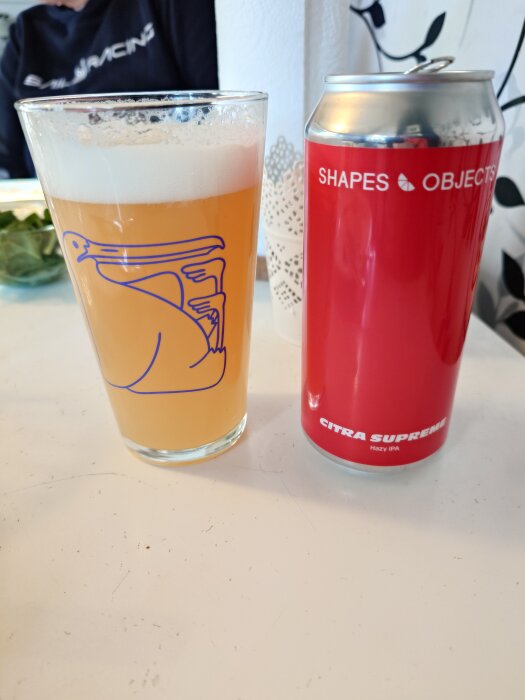 Ett glas med skummande ljus öl och en röd burk märkt "Shapes Objects" och "Citra Supreme Hazy IPA" står på ett bord.