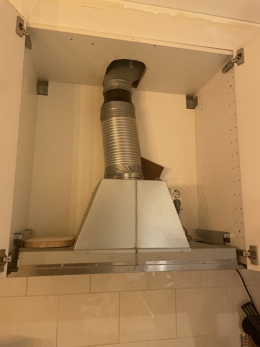 IKEA köksfläkt monterad med utsugsrör i ett skåp, synlig ventilationstejp och ovanliggande eluttag; behöver ändras för kolfilterinstallation.