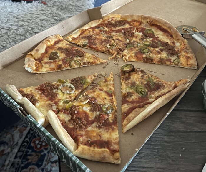 Pizza i kartong med jalapeños och köttfärs som pålägg, skuren i triangulära bitar på ett bord.
