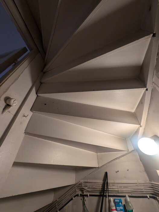 Vy underifrån av en vitmålad trappa med synliga stödstrukturer och rördragningar, bar glödlampa lyser trappan.