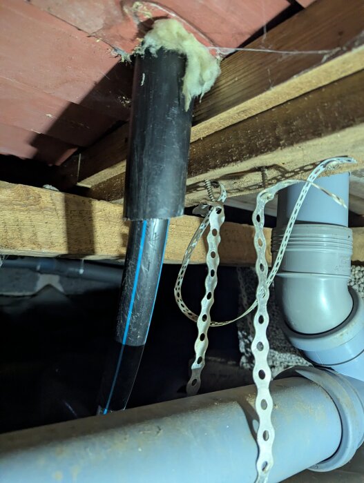 PEM-slang och grått avloppsrör i en krypgrund, vid träbjälklag och isolering. En metallhylsa håller rören på plats.