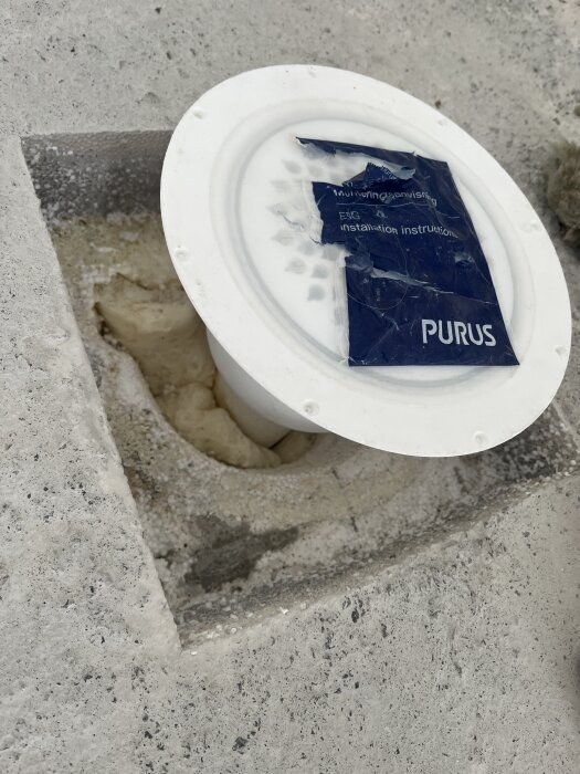 Golvbrunn installerad i ett rum med betonggolv, omgiven av borttagen frigolit. Skyddskåpa med instruktioner från Purus.
