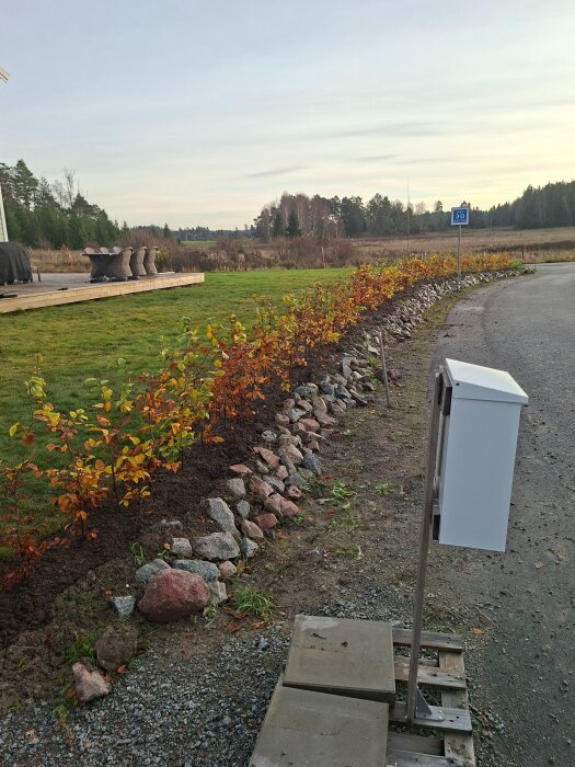 Slänt med stenläggning och nyplanterad häck, omgiven av gräsmatta och väg, betongplattor och skylt längst ner på bilden.