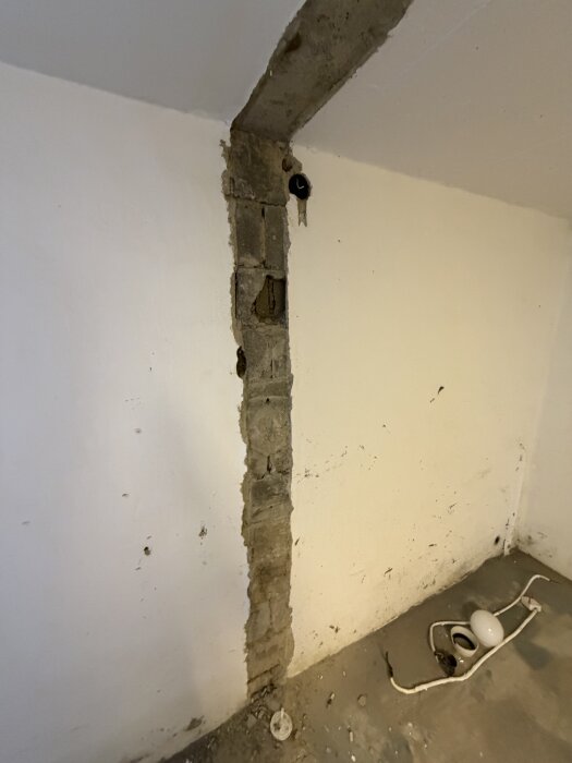 Skadad vägg i källare med synlig puts och eluttag, där djupare sprickor behöver lagas; vitmålad yta med rör och kablar på golv.