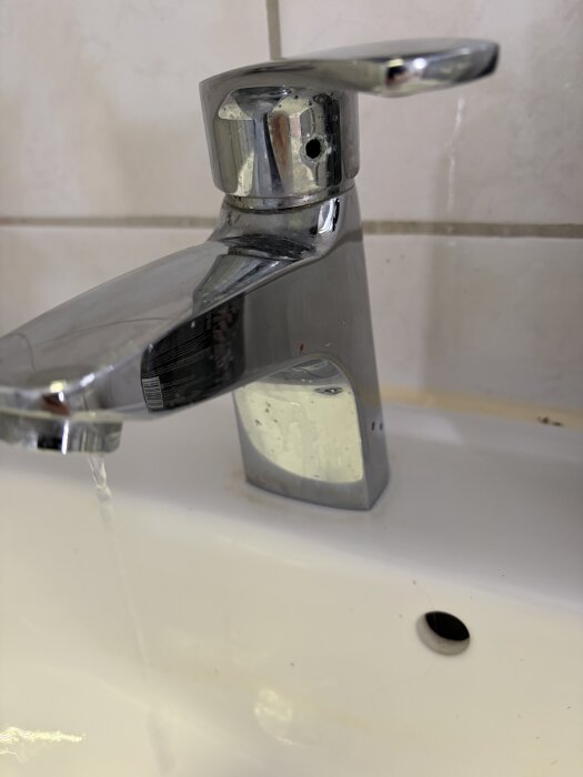 Kromad badrumskran med läckage, vatten droppar från pipen. Kranens bas och handfat synliga, samt del av kakelvägg i bakgrunden.