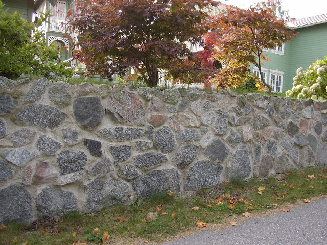 Mur av natursten med murbruk, omgiven av träd och grönska framför ett grönmålat hus.