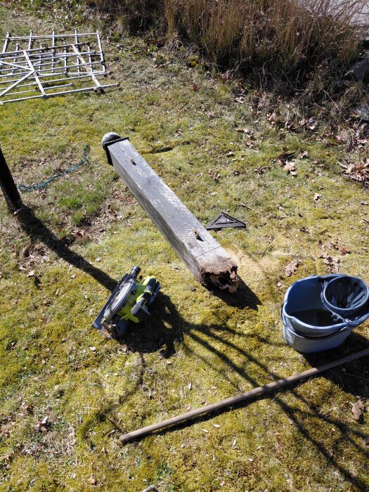 Grindstolpe som har ruttnat vid basen ligger på gräsmatta med verktyg och spann bredvid, omgiven av löv.