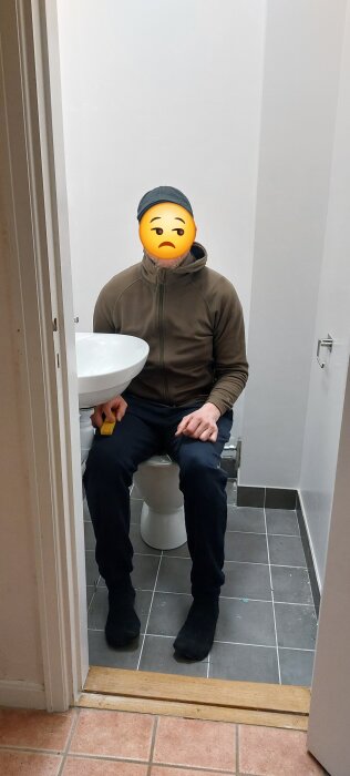Person sitter på en toalettstol i ett litet badrum, hållande ett måttband, med handfat bredvid. Golvet är kaklat och väggarna är ljusa.