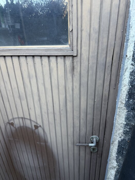 En trädörr med spårpanel, sänkt i övre hörnet, har ett fönster och enkel dörrklinka. Ytterbeklädnaden visar tecken på slitage.