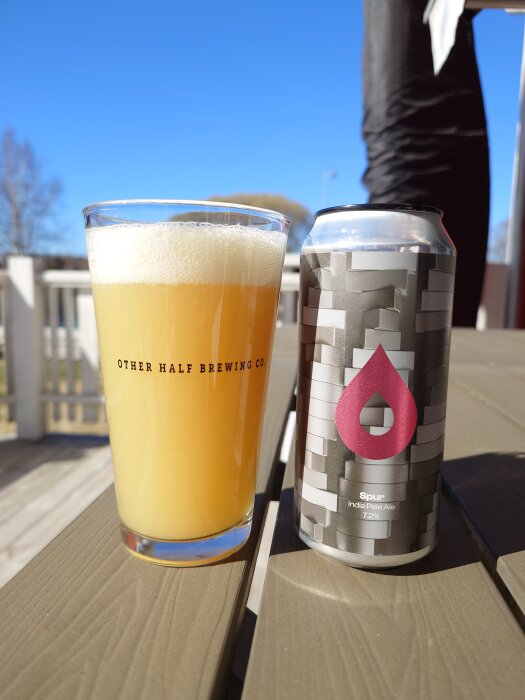 En pintglas med ljus öl från Other Half Brewing Co. bredvid en burk av Spur India Pale Ale står på ett bord mot klarblå himmel.