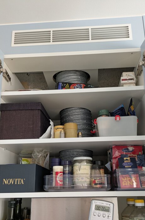 Skåp med flera hyllor fyllda med hushållsartiklar, inklusive kryddor, kex, och runda formar. Plastbehållare och kartonger syns också.
