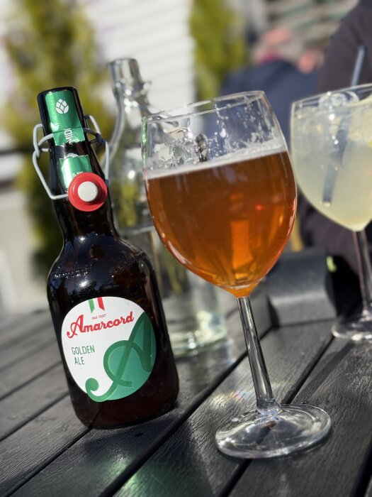 Flaska med Amarcord Golden Ale och ett glas öl står på ett svart utomhusbord i solljus.