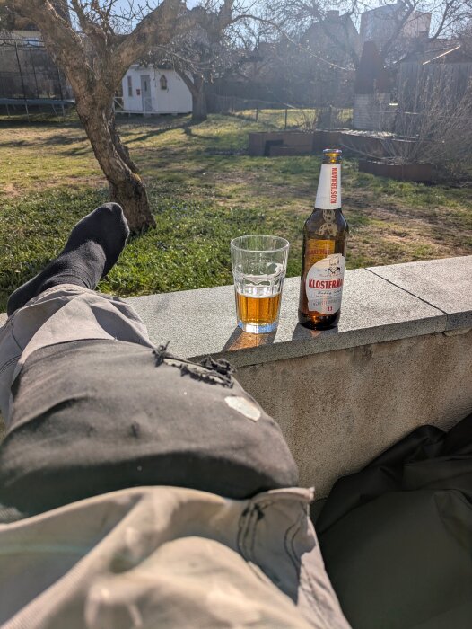 En person ligger ner utomhus, med ett glas öl och en ölflaska på en mur i solljuset.