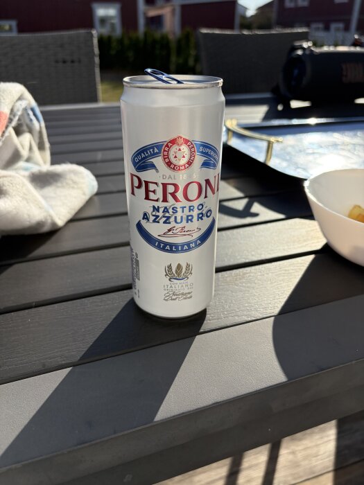 En burk Peroni-öl står på ett trädgårdsbord, omgiven av en trasa och serveringsfat i solljuset.