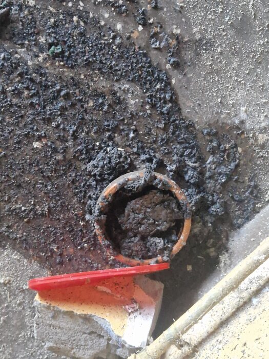 En avloppsledning fylld med smuts och skräp samt en röd plastpinne ligger på ett betonggolv.