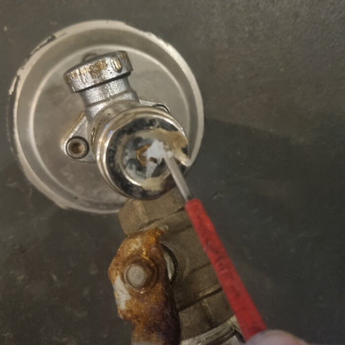 Närbild på en metallventil där en person skrapar med ett verktyg. Ventilen har rostiga delar och sitter på en mörk vägg.