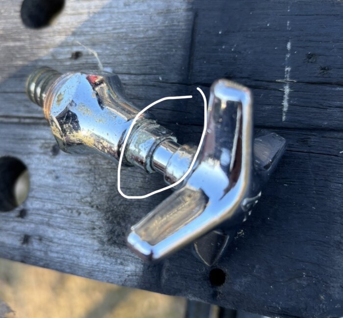 Äldre vattenkran med löst vred, cirkel markerar området runt stången där vattenläckage uppstår. Kranen vilar på ett träbord.