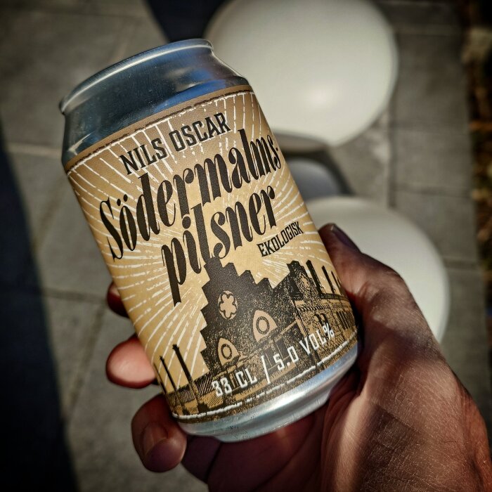 En hand som håller en burk av Nils Oscar Södermalms Pilsner, med en design i bruna toner och texten "Ekologisk".