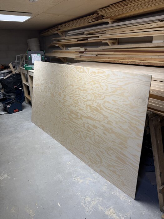 Verkstad med staplade träskivor och en stor plywoodskiva lutad mot en arbetsbänk, omgiven av verktyg och material.