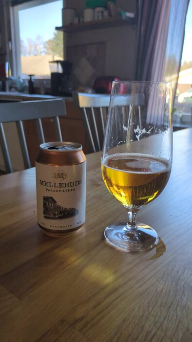 En öppen burk Melleruds Pilsner och ett glas med öl på ett köksbord, med ett fönster i bakgrunden.