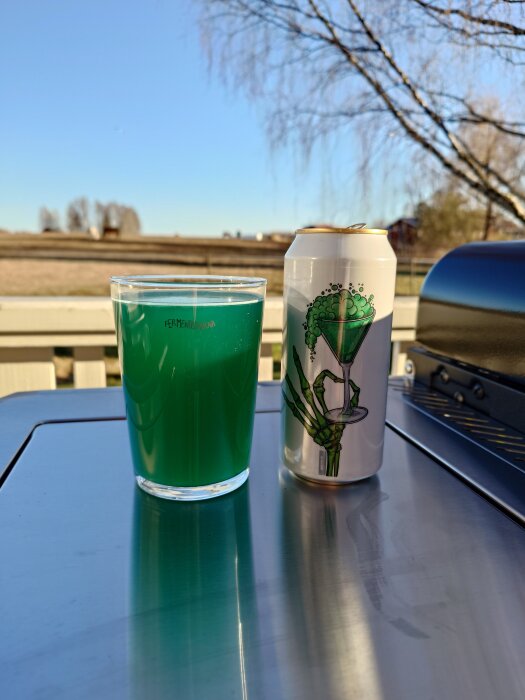 Glas med grönt öl och en burk med kreativ etikett på ett bord utomhus, med grill och träd i bakgrunden under klar himmel.