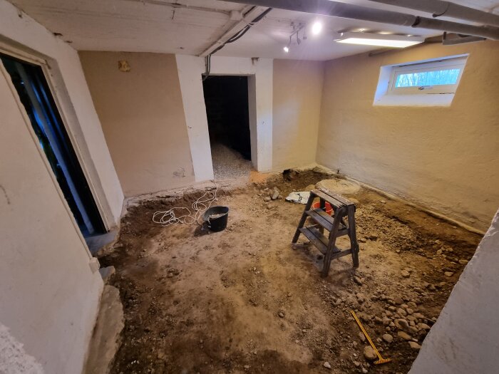 Bild av en källare under renovering med uppbrutet betonggolv, en stege, en hink och synliga rör i taket. Ventilation och naturbelysning från ett fönster.