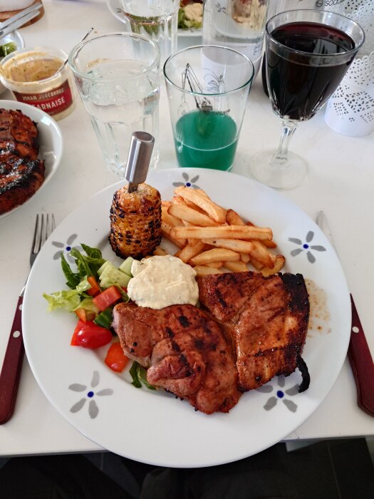 Grillad flintastek med pommes frites, majs, sallad och sås på en tallrik, omgiven av glas med dryck och en burk bearnaisesås.