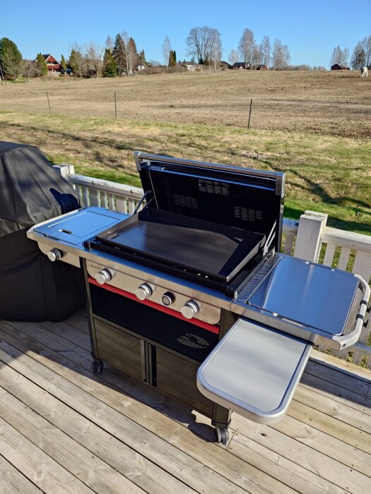 Ny gasolgrill på altan, modern design med fyra brännare och rostfria ytor. Klarblå himmel och landsbygd i bakgrunden. Perfekt för sommarkvällar.