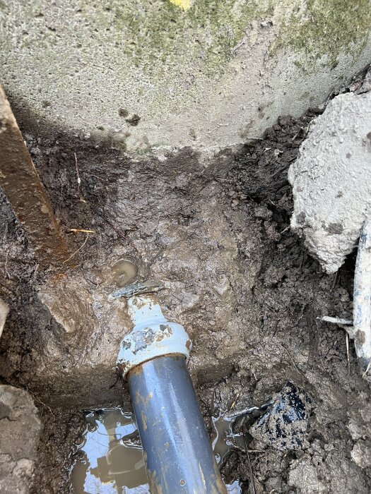 Plaströr med metallkoppling i mark, synligt läckage vid skarven, omgiven av lera och vatten. Kopplingen är delvis ingjuten i en gammal betongbrunn.