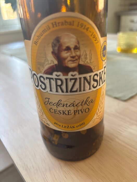 En flaska Postřižinské tjeckiskt öl med etikett som visar en äldre man, namn och ölets styrka, placerad på ett bord.