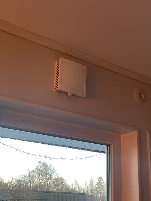 Ventilationsgaller monterat ovanför ett fönster i ett rum med utsikt över träd och byggnader utomhus.