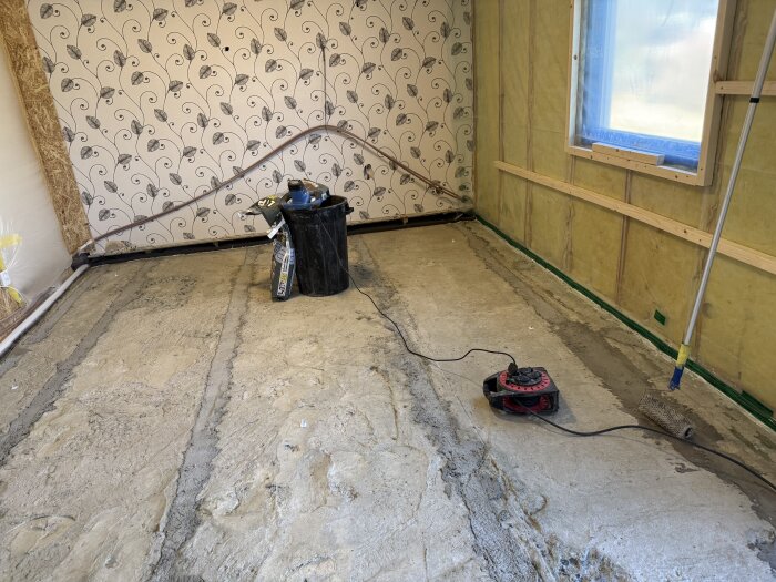 Rum under renovering med nylagd flytspackel, en handvisp och en hink står på golvet, tapetserad vägg med mönster i bakgrunden.