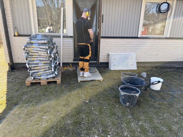 Person står vid hög med säckar med byggmaterial utanför hus, flera hinkar och en slang syns på marken.