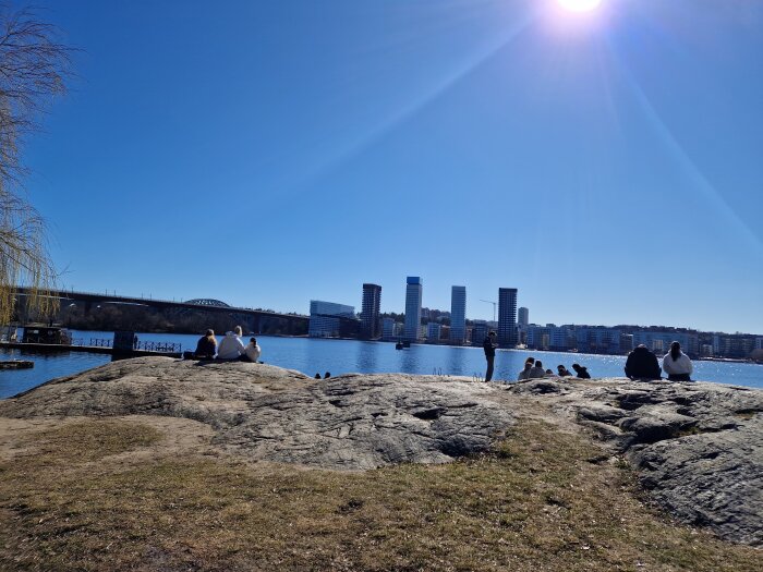 Stadssilhuett över vatten med några personer på klippa i förgrunden och bro i bakgrunden under en klar blå himmel.