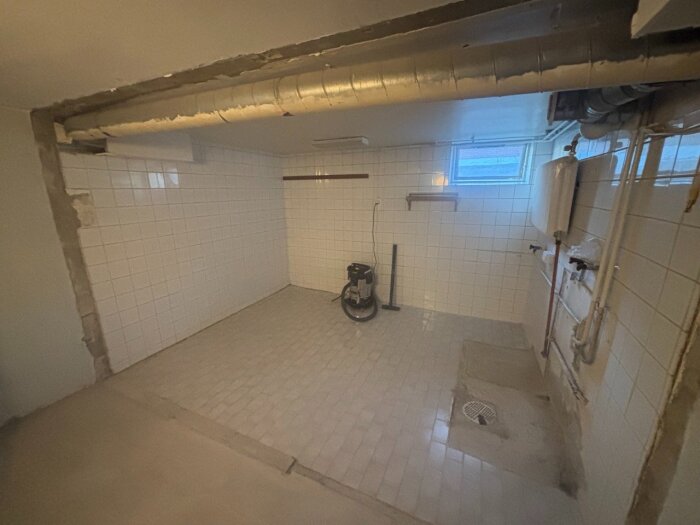 Källare med vitkaklade väggar, rörinstallationer och en dammsugare på klinkergolv, samt ett litet fönster nära taket.