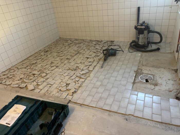Renovering av badrum; delvis borttaget kakelgolv, synliga trasiga plattor och verktyg på golvet, och en våtdammsugare i hörnet.