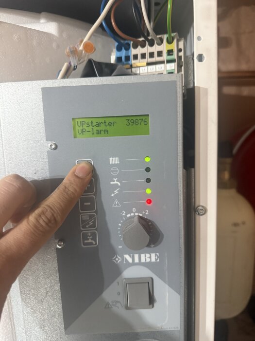 Display och knappar på en NIBE värmepump med indikatorlampor; en hand trycker på en knapp och skärmen visar "VPstarter 39876" och "VP-larm".