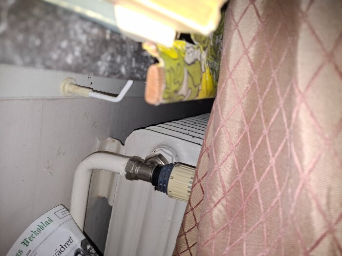 Radiator med gammalt järnrörsanslutet rör synligt bakom ett mönstrat tyg och vid sidan av en tidskrift.